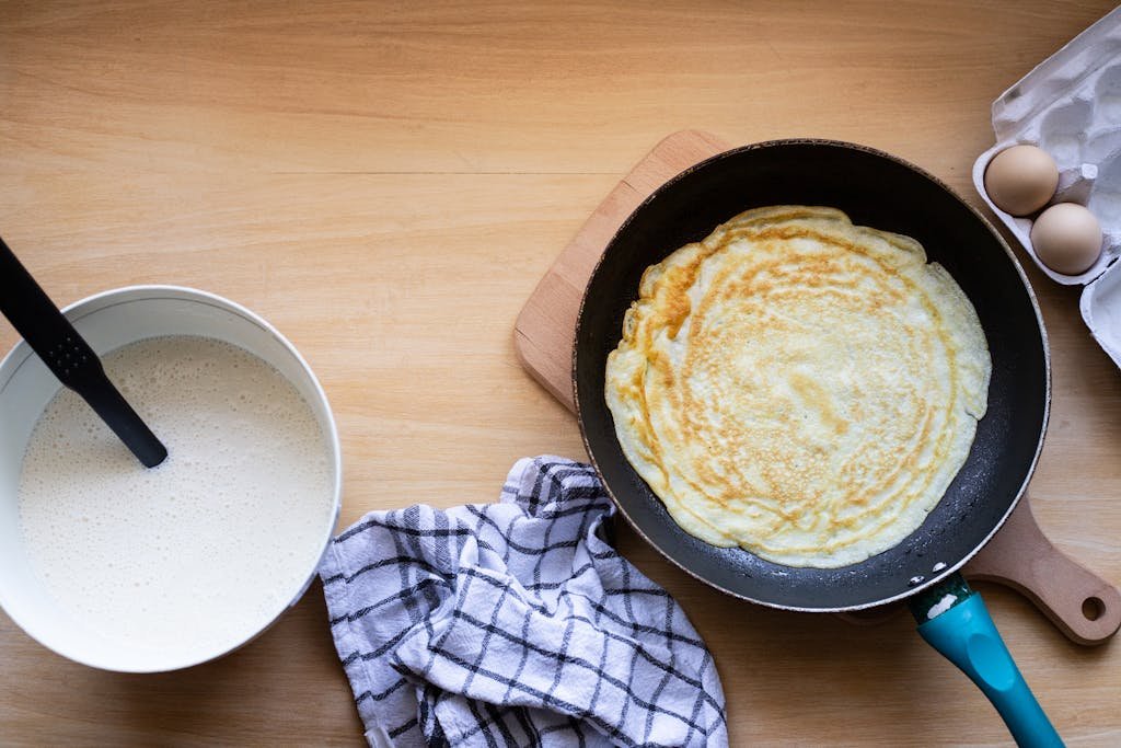 Top view of a pancake cooking in a pan with batter and eggs nearby.